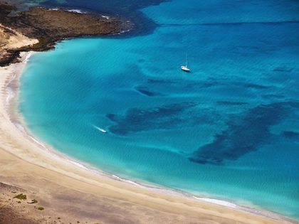 Aerial view of Playa del Risco Lanzarote — wild empty crescent-shaped beach of fine golden sand at the foot of the Risco de Famara cliffs, with crystal-clear turquoise water and volcanic black rock